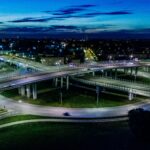 A dramatic cityscape featuring a modern flyover lit by vibrant city lights at night.