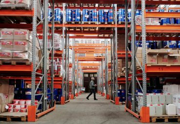 pexels-photo-4483775-4483775 A man walking through a large industrial warehouse with stacked shelves filled with goods and products.